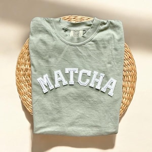 May include: Sage green t-shirt with a white "MATCHA" graphic in a varsity style font.