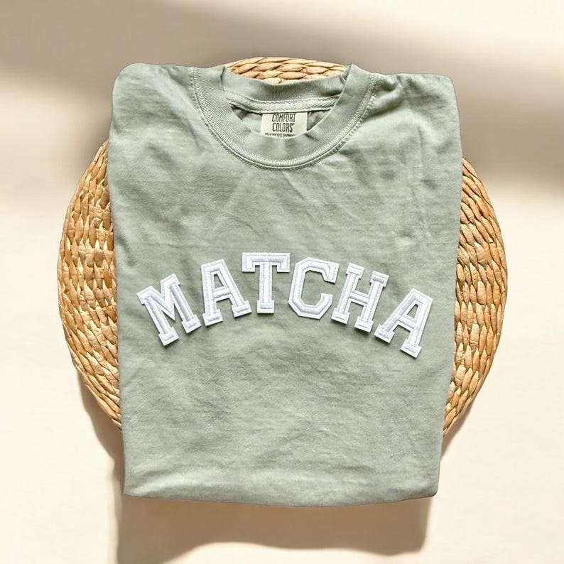 May include: Sage green t-shirt with a white "MATCHA" graphic in a varsity style font.