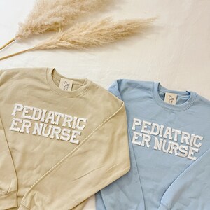 May include: Two sweatshirts with the text "Pediatric ER Nurse" in white block letters. One sweatshirt is light brown and the other is light blue.
