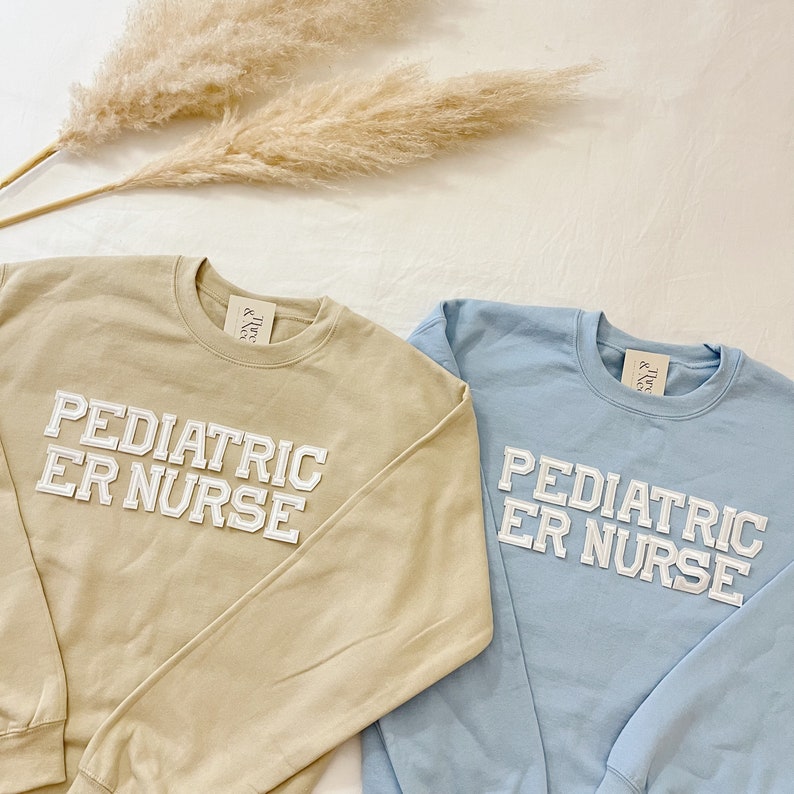 May include: Two sweatshirts with the text "Pediatric ER Nurse" in white block letters. One sweatshirt is light brown and the other is light blue.