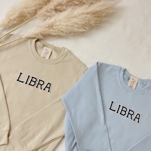 May include: Two sweatshirts with the word "LIBRA" embroidered on the front. One sweatshirt is beige and the other is light blue.