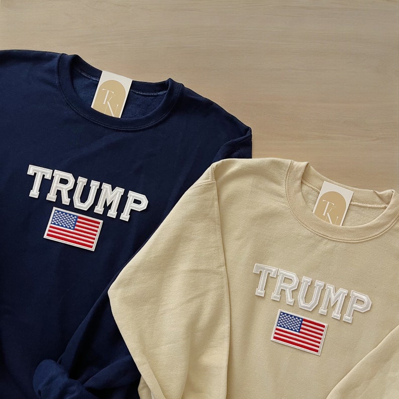 Puede incluir: Dos sudaderas, una azul marino y otra beige, con la palabra "TRUMP" en letras blancas y un parche de la bandera estadounidense.