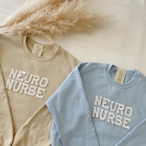 Pu&ograve; includere: Due felpe, una beige e una celeste, con il testo "NEURO NURSE" in lettere bianche a blocchi.