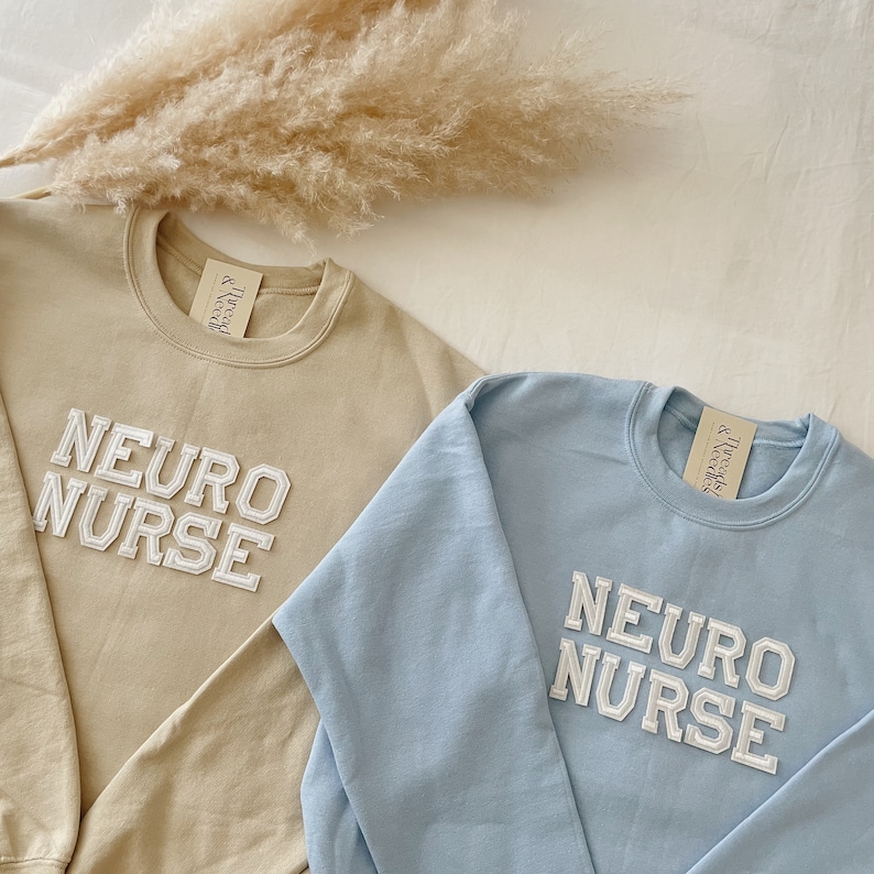 Pu&ograve; includere: Due felpe, una beige e una celeste, con il testo "NEURO NURSE" in lettere bianche a blocchi.