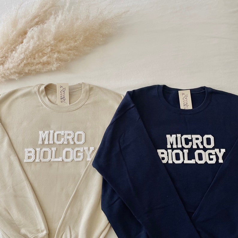 May include: Two crewneck sweatshirts, one beige and one navy blue, each with the text "MICRO BIOLOGY" in white, collegiate-style lettering. The sweatshirts are displayed on a white surface with a fluffy, beige decorative element.