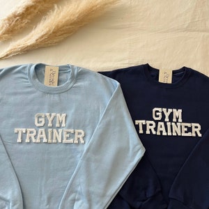 Könnte beinhalten: Zwei langärmelige Sweatshirts mit dem Text "Gym Trainer" in weißen Buchstaben auf blauem und marineblauem Hintergrund.