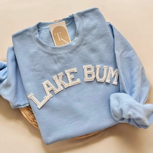 May include: A light blue sweatshirt with the text "LAKE BUM" in white, block-style lettering. The sweatshirt has a crew neck and long sleeves. A small tag is attached near the neckline. The sweatshirt is folded on a woven mat.
