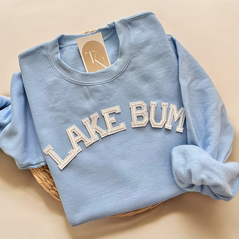 May include: A light blue sweatshirt with the text "LAKE BUM" in white, block-style lettering. The sweatshirt has a crew neck and long sleeves. A small tag is attached near the neckline. The sweatshirt is folded on a woven mat.