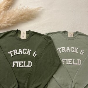 Könnte beinhalten: Zwei langärmelige Sweatshirts in Olivgrün und Hellgrün mit dem Text "TRACK & FIELD" in weißen umrandeten Buchstaben.