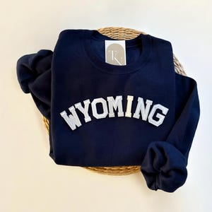 Könnte beinhalten: Ein marineblauer Sweatshirt mit einem weißen Filz-Applikationen des Wortes "WYOMING" in einer College-Schrift.