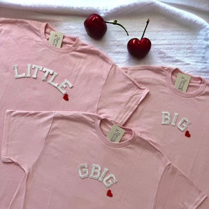May include: Three pink t-shirts with white text. The shirts say "Little" with a red heart, "Big" with a red heart, and "GBIG" with a red heart. The shirts are on a white surface with red cherries.