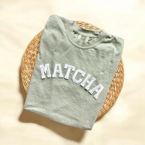 May include: A light green t-shirt with the word "MATCHA" in white block letters on the front. The shirt is on a woven straw placemat.