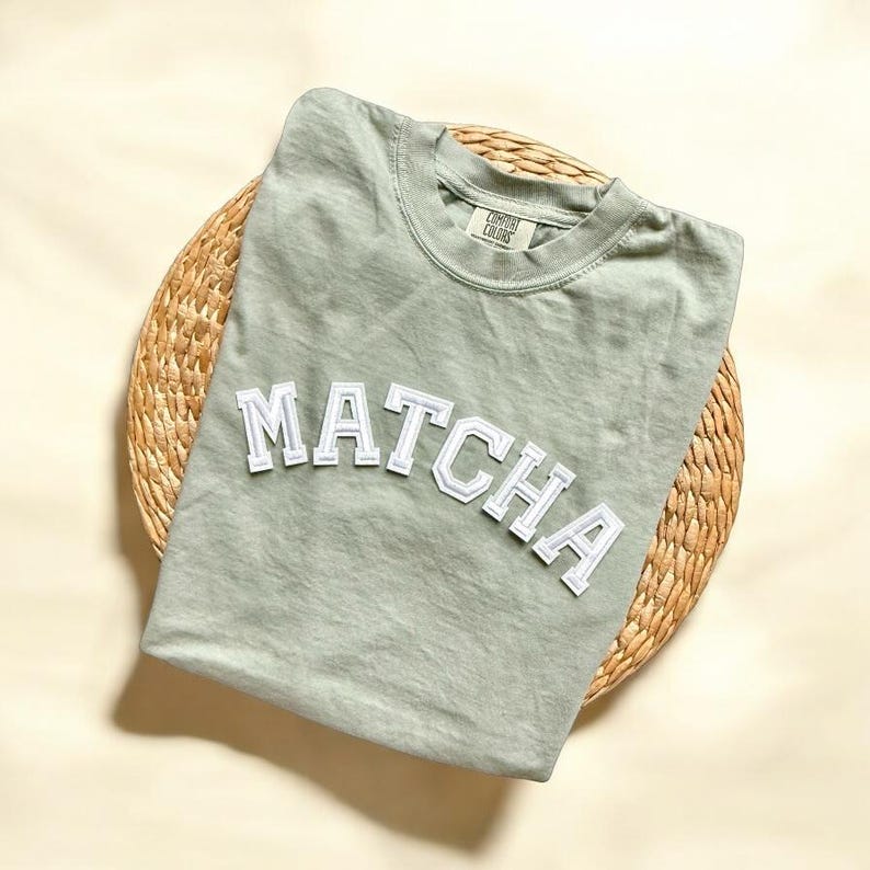 May include: A light green t-shirt with the word "MATCHA" in white block letters on the front. The shirt is on a woven straw placemat.