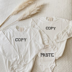 May include: Three white sweatshirts with black text. The largest sweatshirt says "COPY" in large block letters. The medium sweatshirt says "COPY" in large block letters. The smallest sweatshirt says "PASTE" in large block letters.