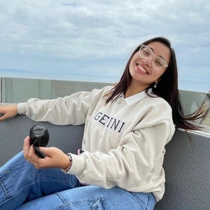May include: A person wearing a light beige sweatshirt with the word "GEINI" embroidered on the front. They are wearing blue jeans and clear framed glasses. They are holding a black object in their hand.