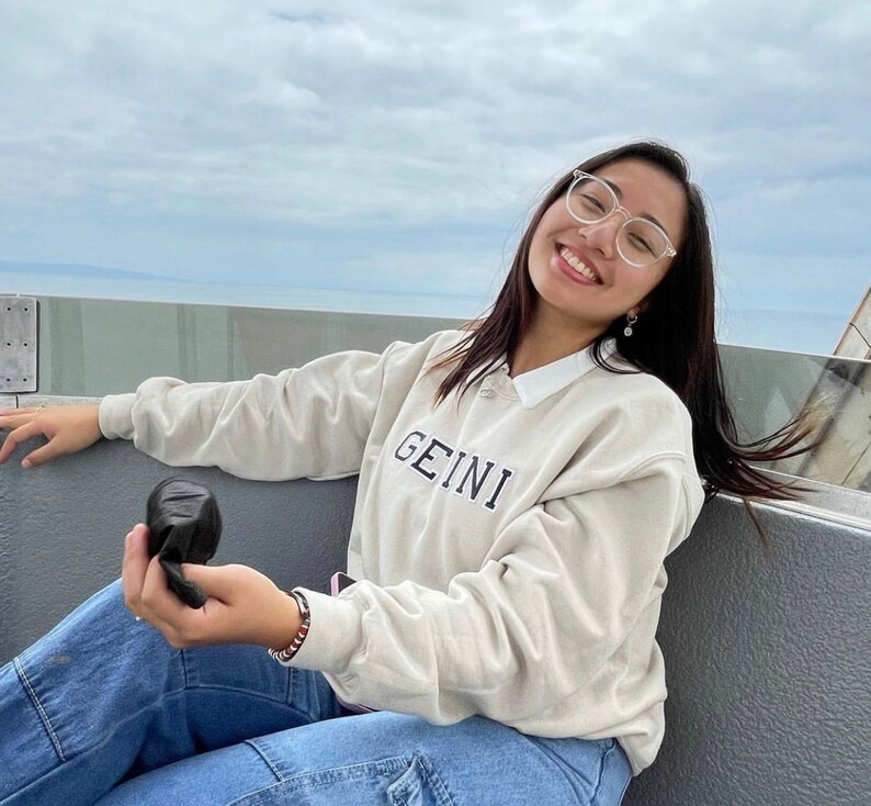 May include: A person wearing a light beige sweatshirt with the word "GEINI" embroidered on the front. They are wearing blue jeans and clear framed glasses. They are holding a black object in their hand.