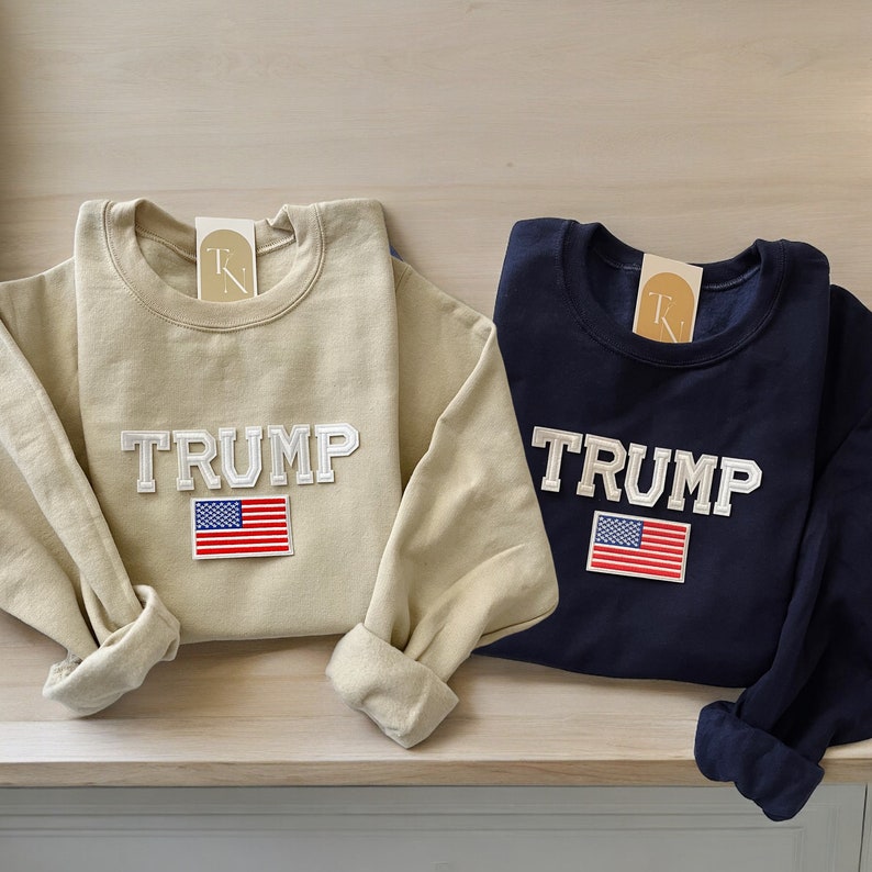 Puede incluir: Dos sudaderas de manga larga con la palabra "TRUMP" y un parche de la bandera estadounidense. Una sudadera es beige y la otra es azul marino.