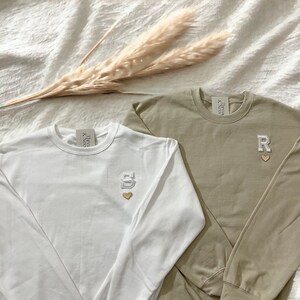 Puede incluir: Dos camisas de manga larga con letras y corazones bordados. La camisa de la izquierda es blanca con una 'S' y la camisa de la derecha es caqui con una 'R'.