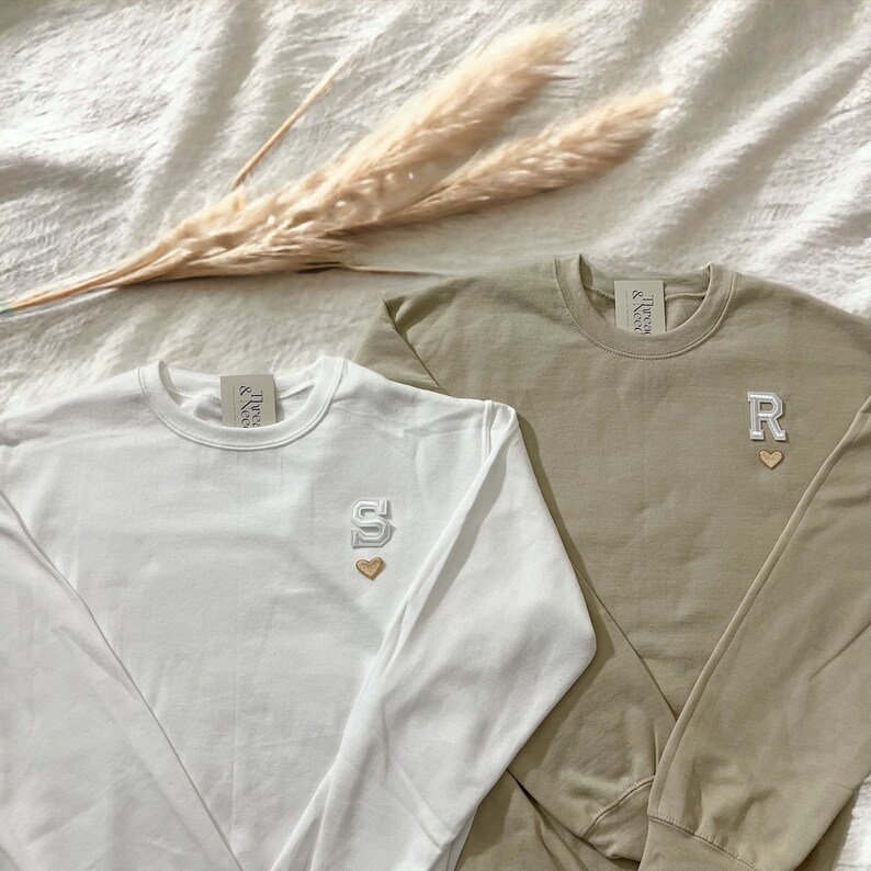 Puede incluir: Dos camisas de manga larga con letras y corazones bordados. La camisa de la izquierda es blanca con una 'S' y la camisa de la derecha es caqui con una 'R'.