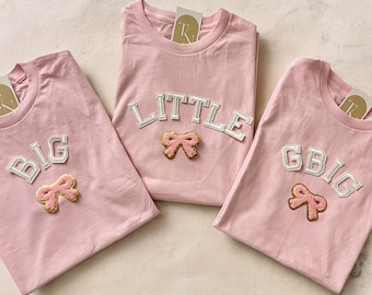 May include: Three pink t-shirts with white lettering and a pink bow applique. The shirts read "BIG", "LITTLE", and "GBIG". Each shirt has a tag attached at the neckline.
