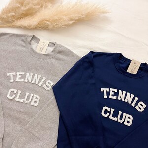 May include: Two long-sleeved sweatshirts with the text "Tennis Club" in white letters on the front. One sweatshirt is grey and the other is navy blue.