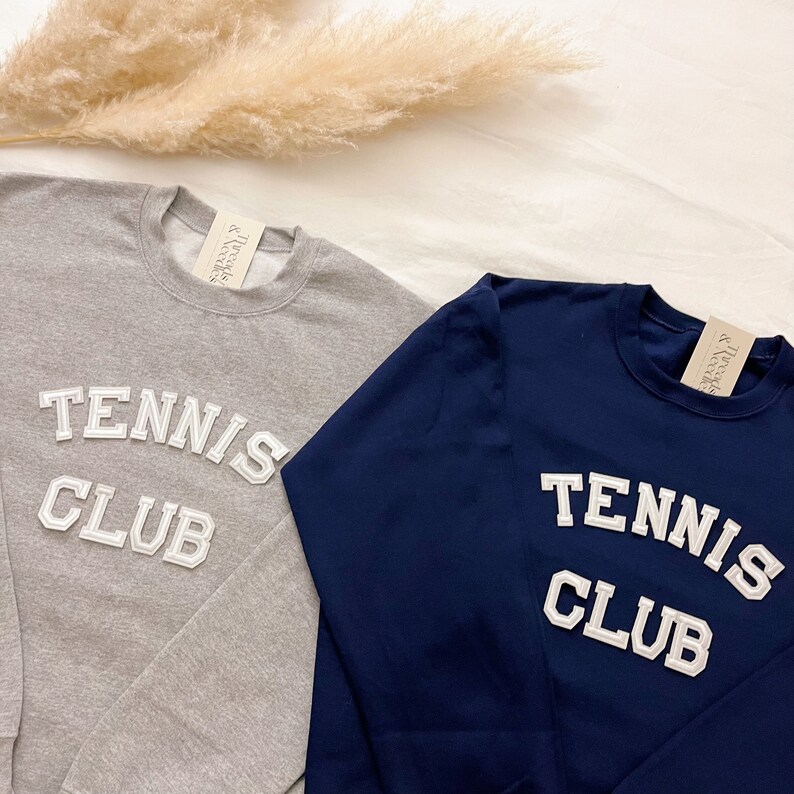 May include: Two long-sleeved sweatshirts with the text "Tennis Club" in white letters on the front. One sweatshirt is grey and the other is navy blue.