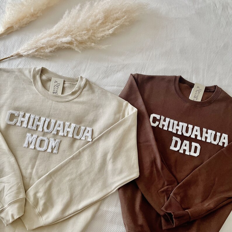 May include: Two crewneck sweatshirts, one tan and one brown, with the words "Chihuahua Mom" and "Chihuahua Dad" in white, block lettering. The sweatshirts are displayed on a white surface with dried ornamental grass.