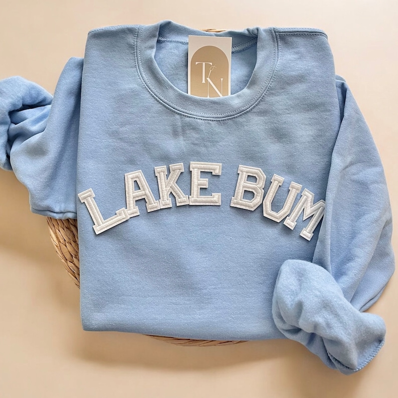 May include: A light blue sweatshirt with the text "LAKE BUM" in white, block-style lettering. The sweatshirt is folded on a woven mat, with a small tag tucked into the collar. The sleeves are folded to the side.