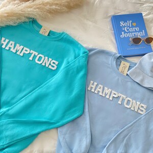 May include: Two long-sleeved sweatshirts with the word "HAMPTONS" in white letters on the front. One sweatshirt is turquoise blue and the other is light blue.