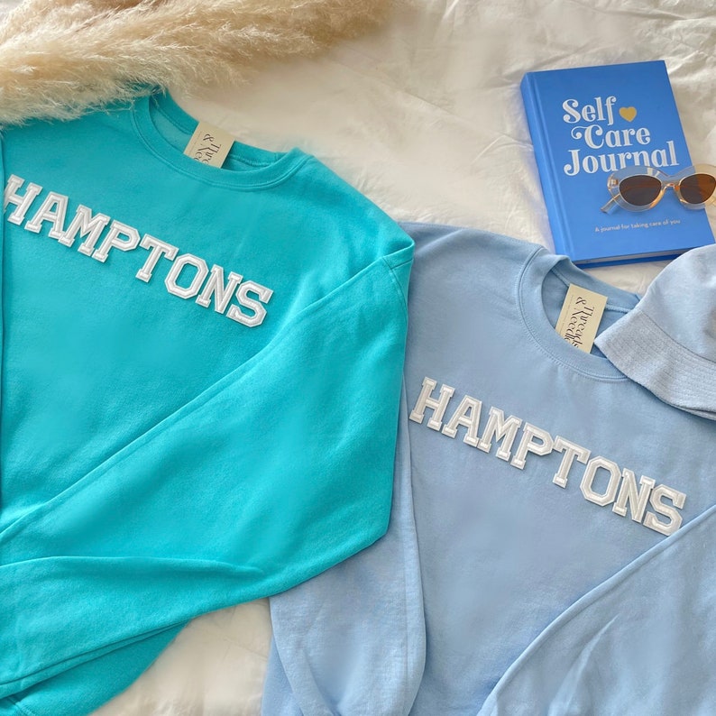 May include: Two long-sleeved sweatshirts with the word "HAMPTONS" in white letters on the front. One sweatshirt is turquoise blue and the other is light blue.