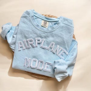 K&ouml;nnte beinhalten: Ein hellblauer Langarmsweatshirt mit den Worten "AIRPLANE MODE" in wei&szlig;en umrandeten Buchstaben.