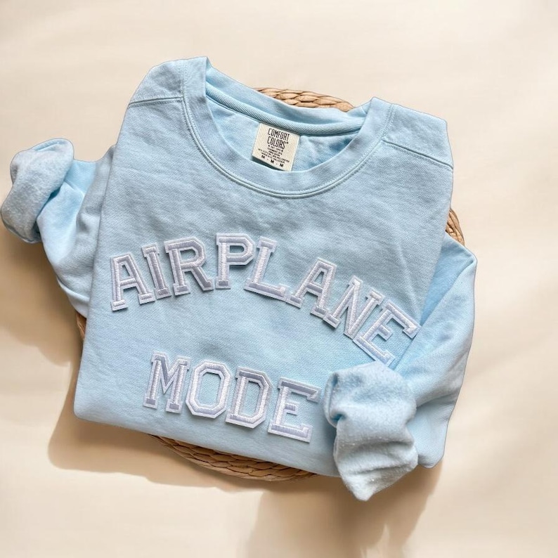K&ouml;nnte beinhalten: Ein hellblauer Langarmsweatshirt mit den Worten "AIRPLANE MODE" in wei&szlig;en umrandeten Buchstaben.