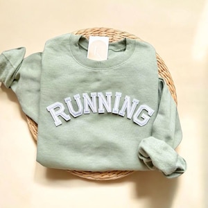 Könnte beinhalten: Ein salbeigrüner Pullover mit dem Wort "RUNNING" in weißen Filzbuchstaben.