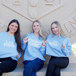 Pu&ograve; includere: Tre donne indossano felpe di colore azzurro chiaro con testo bianco. Il testo sulle felpe recita "L & D Nurse", "Orthopedic Nurse" e "ICU Nurse". Le donne sono sedute su una panchina di pietra di fronte a un edificio in pietra.