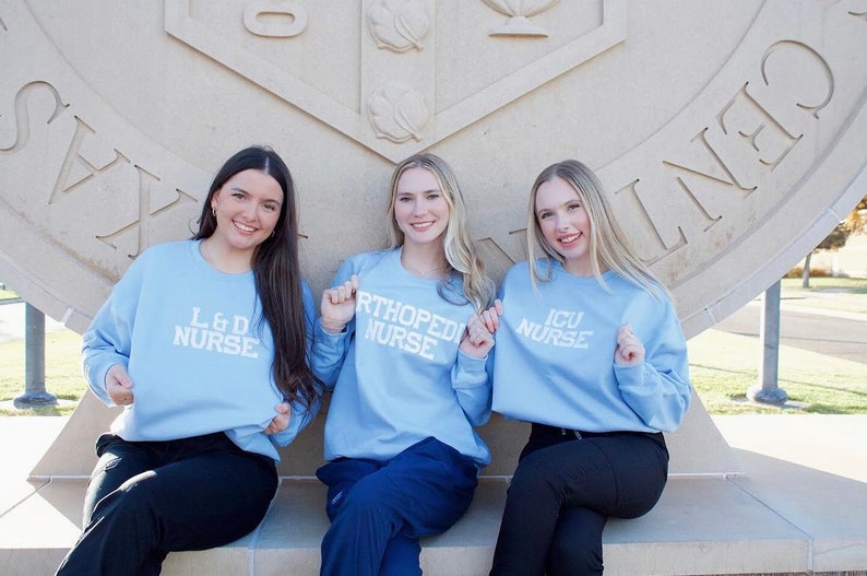Pu&ograve; includere: Tre donne indossano felpe di colore azzurro chiaro con testo bianco. Il testo sulle felpe recita "L & D Nurse", "Orthopedic Nurse" e "ICU Nurse". Le donne sono sedute su una panchina di pietra di fronte a un edificio in pietra.