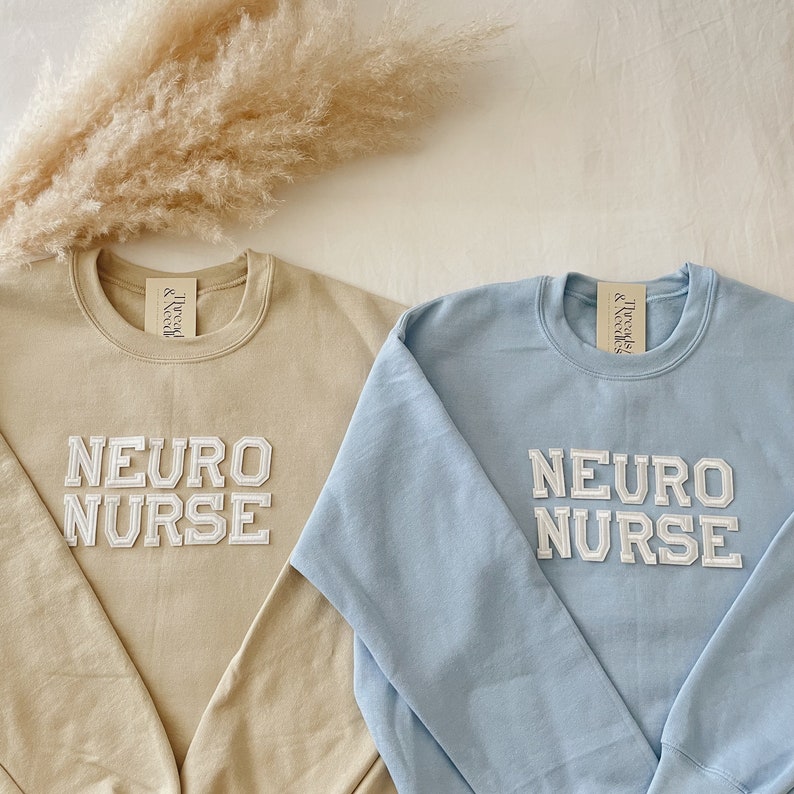 Pu&ograve; includere: Due felpe con il testo "Neuro Nurse" in lettere bianche a blocchi su una felpa beige e una felpa blu chiaro.