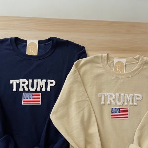 Puede incluir: Dos sudaderas de manga larga con la palabra "TRUMP" en letras blancas y un parche de la bandera estadounidense. Una sudadera es azul marino y la otra es beige.