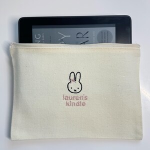Pu&ograve; includere: Una pochette con cerniera in tela bianca con un coniglio ricamato rosa e il testo "lauren's kindle".