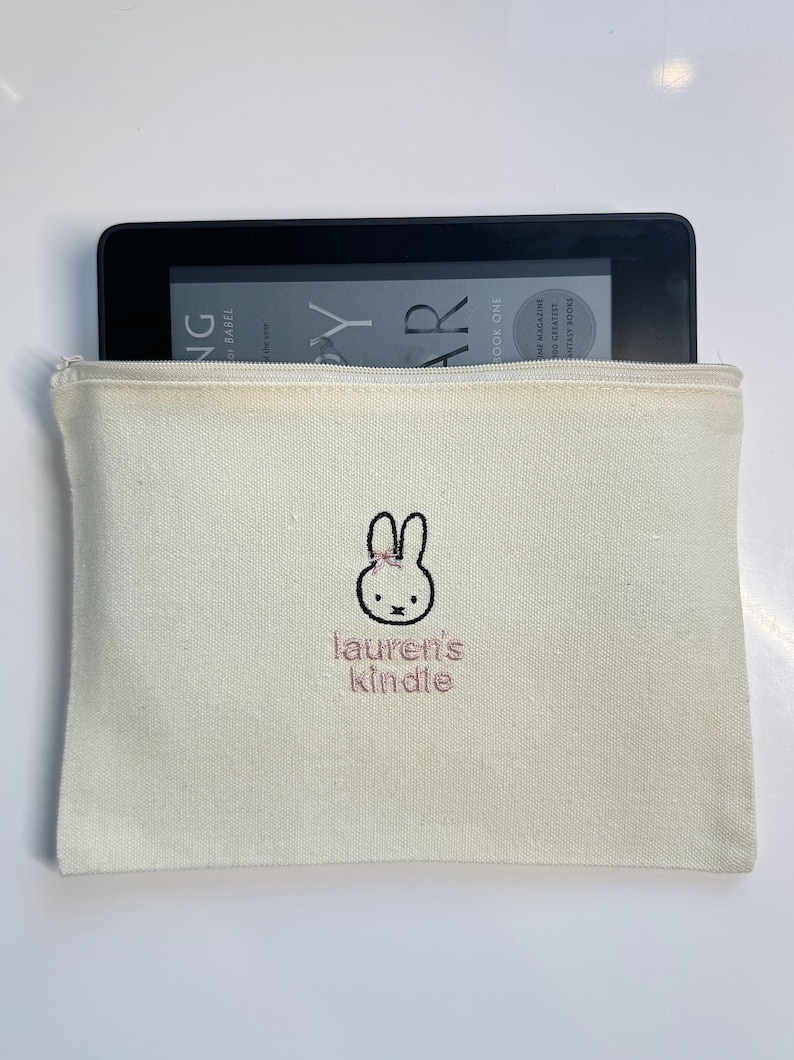 Pu&ograve; includere: Una pochette con cerniera in tela bianca con un coniglio ricamato rosa e il testo "lauren's kindle".