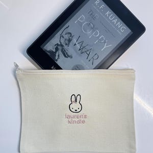 Pu&ograve; includere: Una pochette con cerniera in tela bianca con un coniglio ricamato rosa e il testo "lauren's kindle".