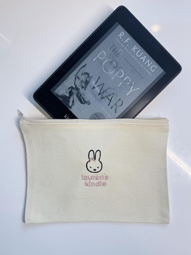 Pu&ograve; includere: Una pochette con cerniera in tela bianca con un coniglio ricamato rosa e il testo "lauren's kindle".