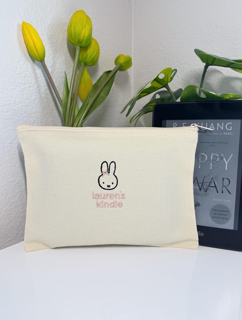 Pu&ograve; includere: Una pochette in tela bianca con un coniglio ricamato rosa e il testo "lauren's kindle".