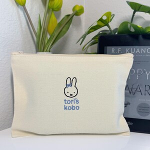 Pu&ograve; includere: Una pochette con cerniera in tela bianca con un coniglio ricamato in blu e il testo "tori's kobo".