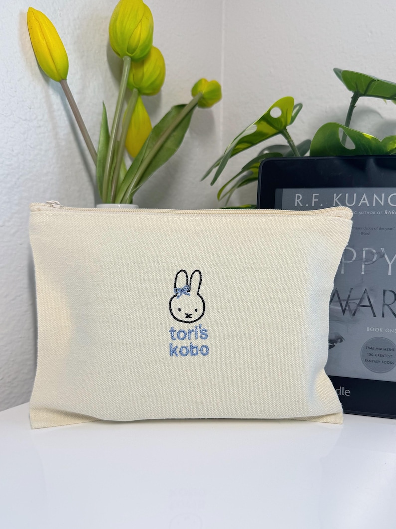 Pu&ograve; includere: Una pochette con cerniera in tela bianca con un coniglio ricamato in blu e il testo "tori's kobo".