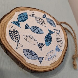 May include: A round wooden ornament with a rope hanger, painted with a white background and blue fish designs.