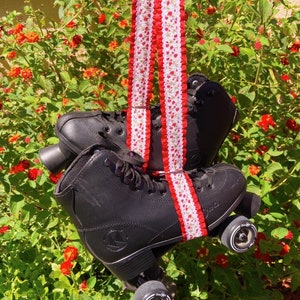 May include: Black roller skates with white wheels and a red and white floral patterned strap. The skates are hanging from the strap in front of a green bush with red flowers.