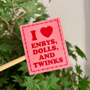 Puede incluir: Pegatina rosa con un borde de alambre de púas. La pegatina dice "I heart Enbys, Dolls, and Twinks" en texto rojo.