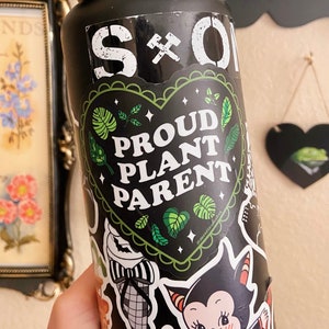 Puede incluir: Botella de agua reutilizable negra con una pegatina verde y negra en forma de corazón que dice "Proud Plant Parent". La botella está cubierta de otras pegatinas.