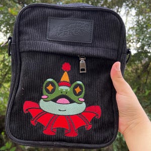 Op de afbeelding: Zwarte corduroy schoudertas met een ritssluiting en een geborduurde kikker met een feestmuts op de voorkant. De tas heeft de tekst "Mister Mister" op de bovenklep.
