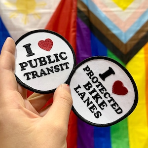 May include: Two round white patches with black borders. One says "I ❤️ PUBLIC TRANSIT" and the other says "I ❤️ PROTECTED BIKE LANES". The heart is red. The background includes flags with rainbow and other colors.